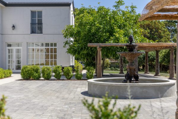 Elegant two-story coastal European-style building with a central fountain, pergola seating, and forest backdrop