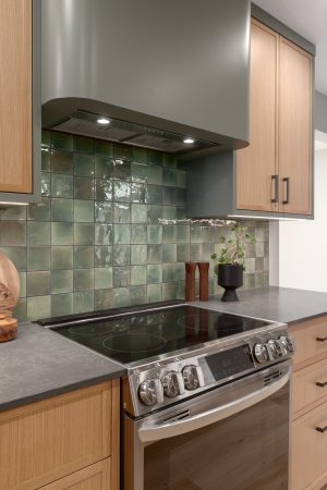 Modern kitchen renovation with stainless steel range, green tile backsplash, and custom wood cabinetry.