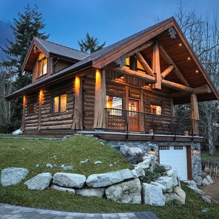 Cozy log cabin with stone foundation, warm exterior lighting, and surrounding trees in Qualicum Beach built by Pickard Construction, Summit Log & Timber Homes and designed by Streamline Designs.
